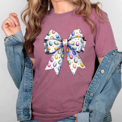 May include: A mauve-colored t-shirt featuring a large bow design. The bow is white with colorful Easter eggs and gold stars. The shirt is paired with a denim jacket.
