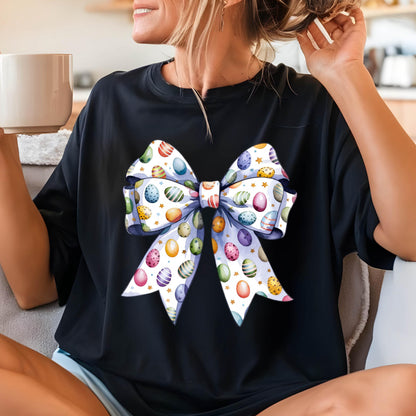 May include: Black t-shirt featuring a large, colorful bow design. The bow is decorated with Easter eggs in various colors, including blue, green, pink, and yellow, with gold stars on a white background. The bow has a light purple border.