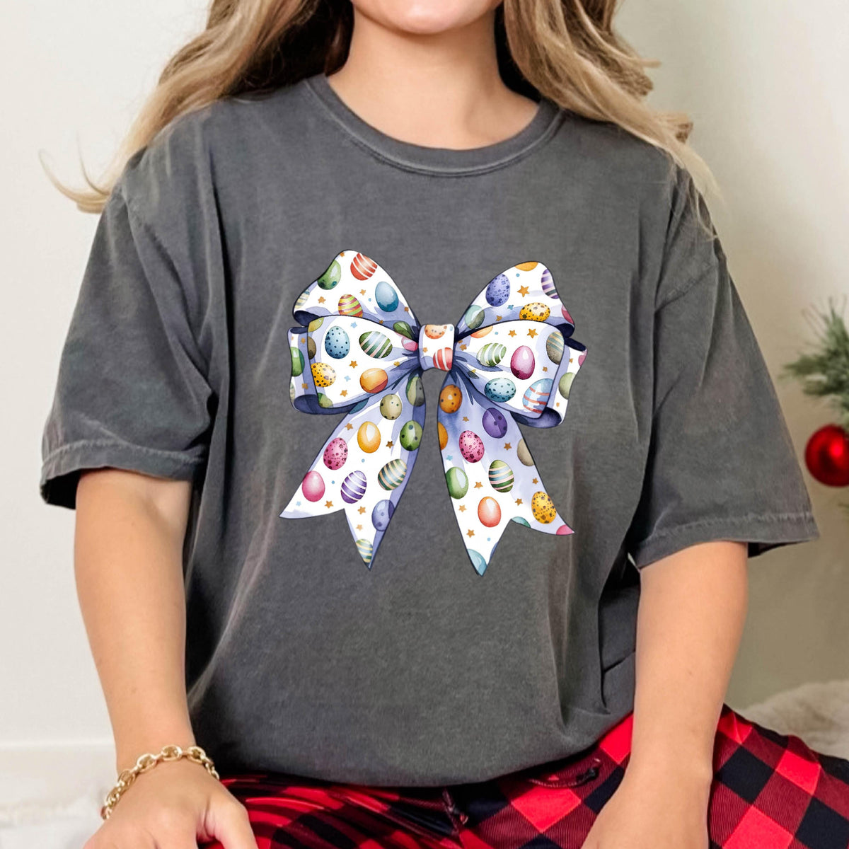 May include: Dark gray t-shirt featuring a large bow design with colorful Easter eggs. The bow has a white background with blue, purple, yellow, and orange eggs. Gold stars accent the design, creating a festive look.