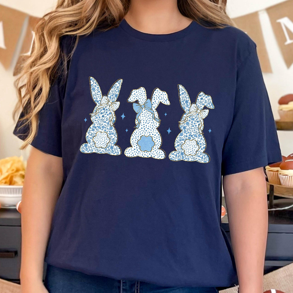 May include: A navy blue t-shirt with a graphic of three blue and white patterned bunnies with bows. The bunnies are facing away from the viewer.