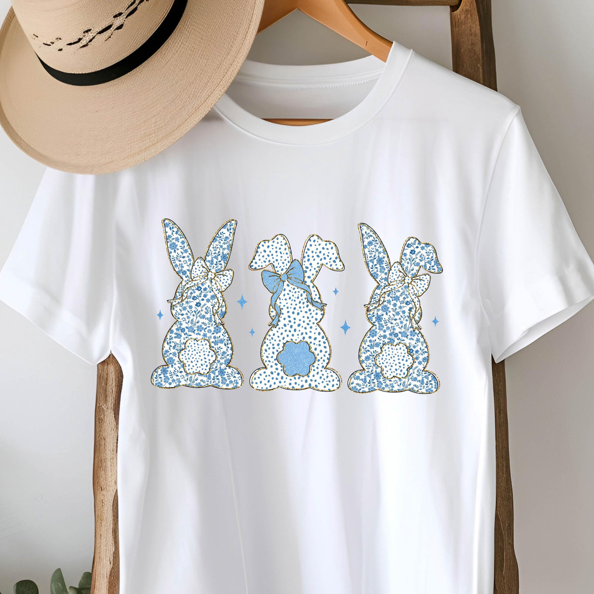 May include: White t-shirt with a blue and gold floral pattern of three bunnies with bows.