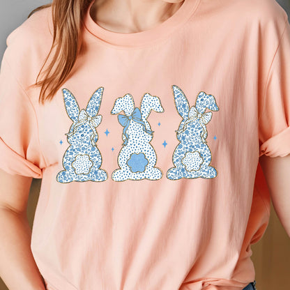 May include: A light pink t-shirt with a graphic of three blue bunnies with white polka dots and blue bows. The bunnies are facing away from the viewer.