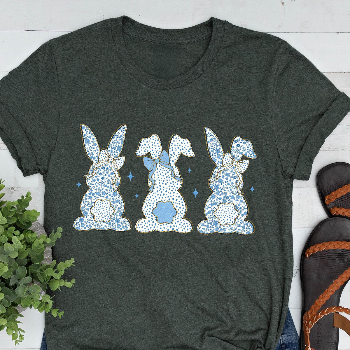 May include: A dark green t-shirt with a graphic of three blue and white bunny rabbits with blue bows. The bunnies are facing away from the viewer and have a white polka dot pattern.