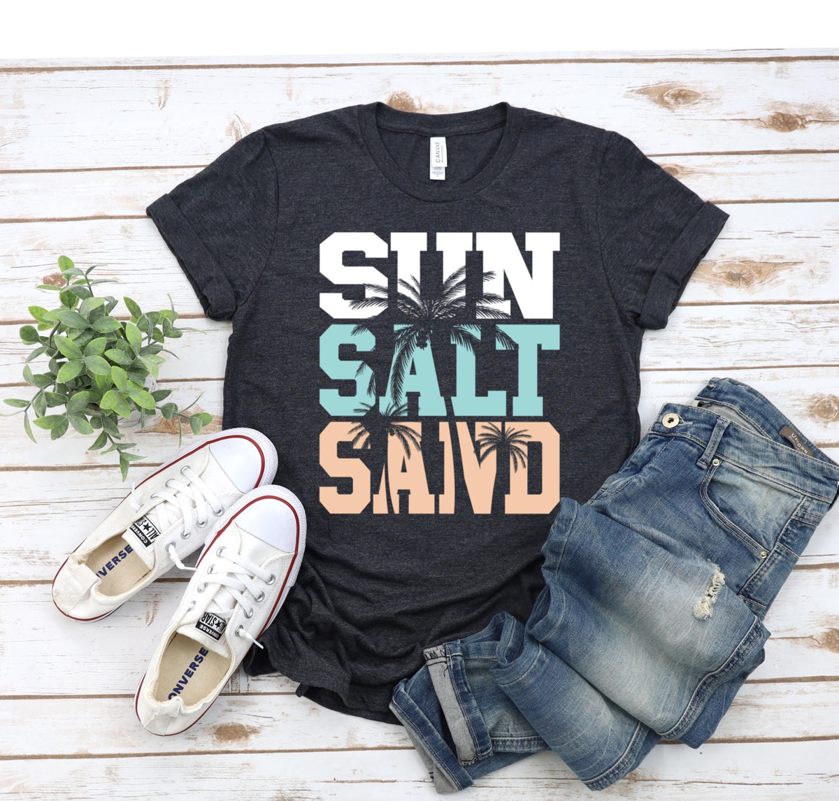May include: A dark gray t-shirt with the words "SUN SALT SAND" in white letters with palm trees in the background. The words are stacked on top of each other with the word "SUN" at the top and the word "SAND" at the bottom. The palm trees are in a light blue and peach color.
