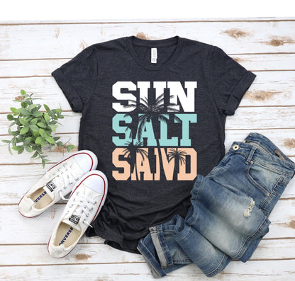 May include: A dark gray t-shirt with the words "SUN SALT SAND" in white letters with palm trees in the background. The words are stacked on top of each other with the word "SUN" at the top and the word "SAND" at the bottom. The palm trees are in a light blue and peach color.
