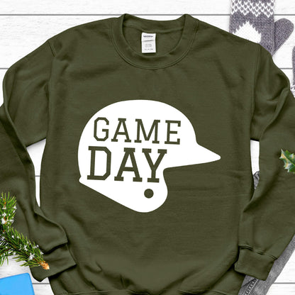 May include: An olive green long-sleeve crewneck sweatshirt featuring a large white graphic of a baseball helmet silhouette. The text "GAME DAY" is printed in a bold, dark olive green block font across the center of the helmet graphic.