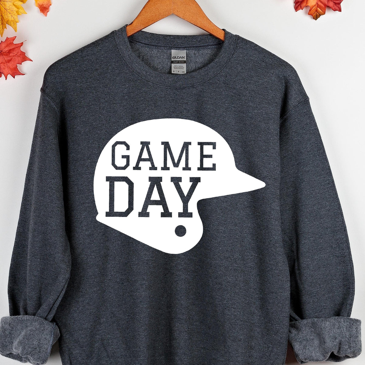 May include: A dark heather gray long-sleeve crewneck sweatshirt featuring a large white graphic of a baseball helmet with the text "GAME DAY" printed in a bold, dark gray block font across the center of the helmet shape on the front.