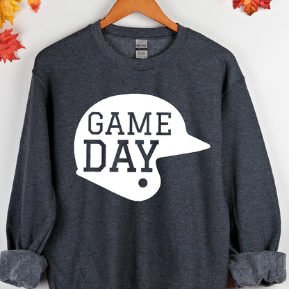 May include: A dark heather gray long-sleeve crewneck sweatshirt featuring a large white graphic of a baseball helmet with the text "GAME DAY" printed in a bold, dark gray block font across the center of the helmet shape on the front.