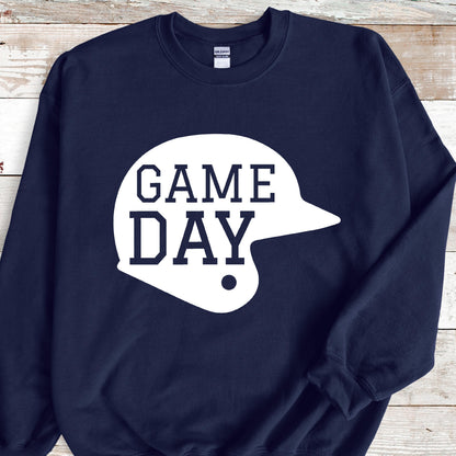 May include: A navy blue crewneck sweatshirt featuring a large white graphic of a baseball helmet silhouette. The text GAME DAY is printed in a bold, dark navy blue font inside the white helmet shape, centered on the front of the garment.