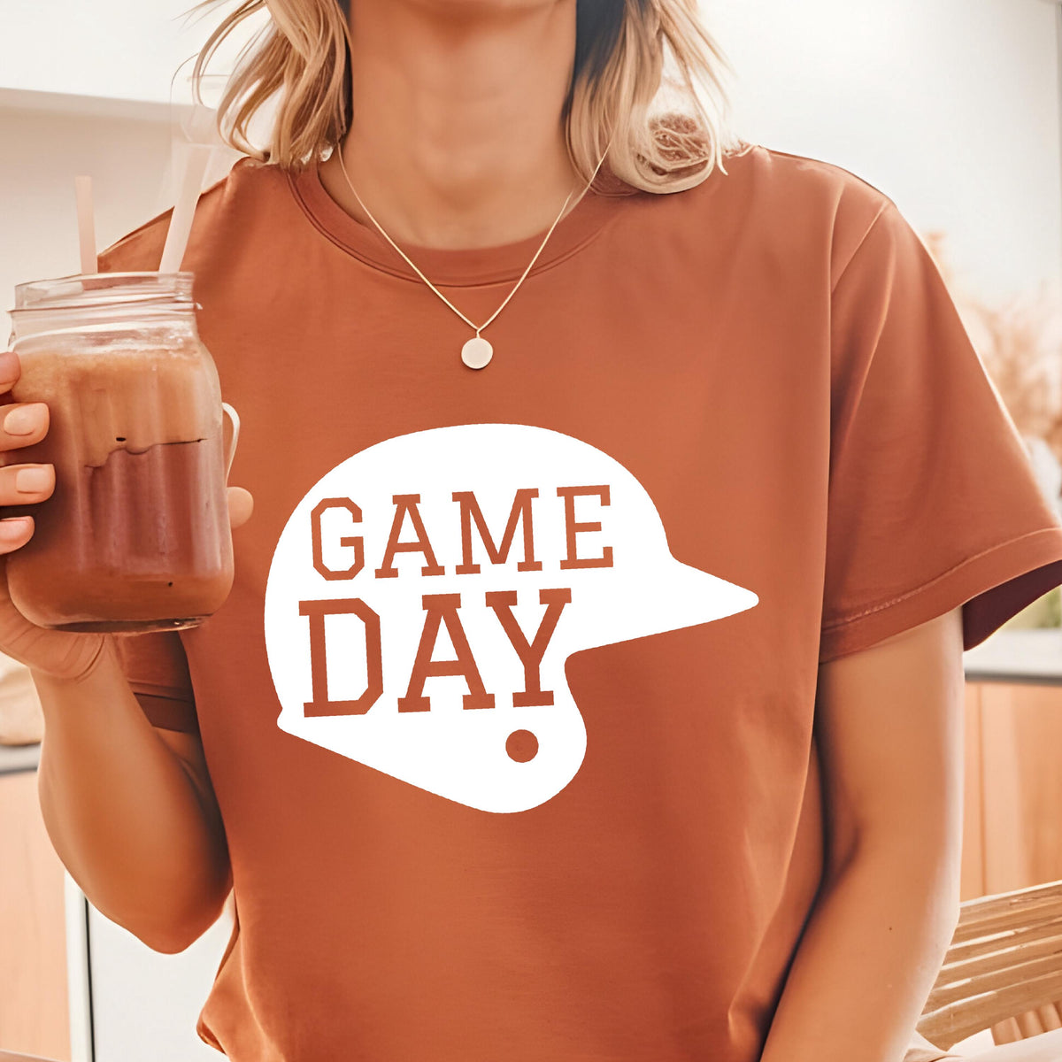 May include: A rust-colored short-sleeve t-shirt featuring a white graphic of a baseball helmet silhouette with the text GAME DAY printed in rust-colored letters inside the helmet shape. The shirt has a crew neckline and a standard fit.