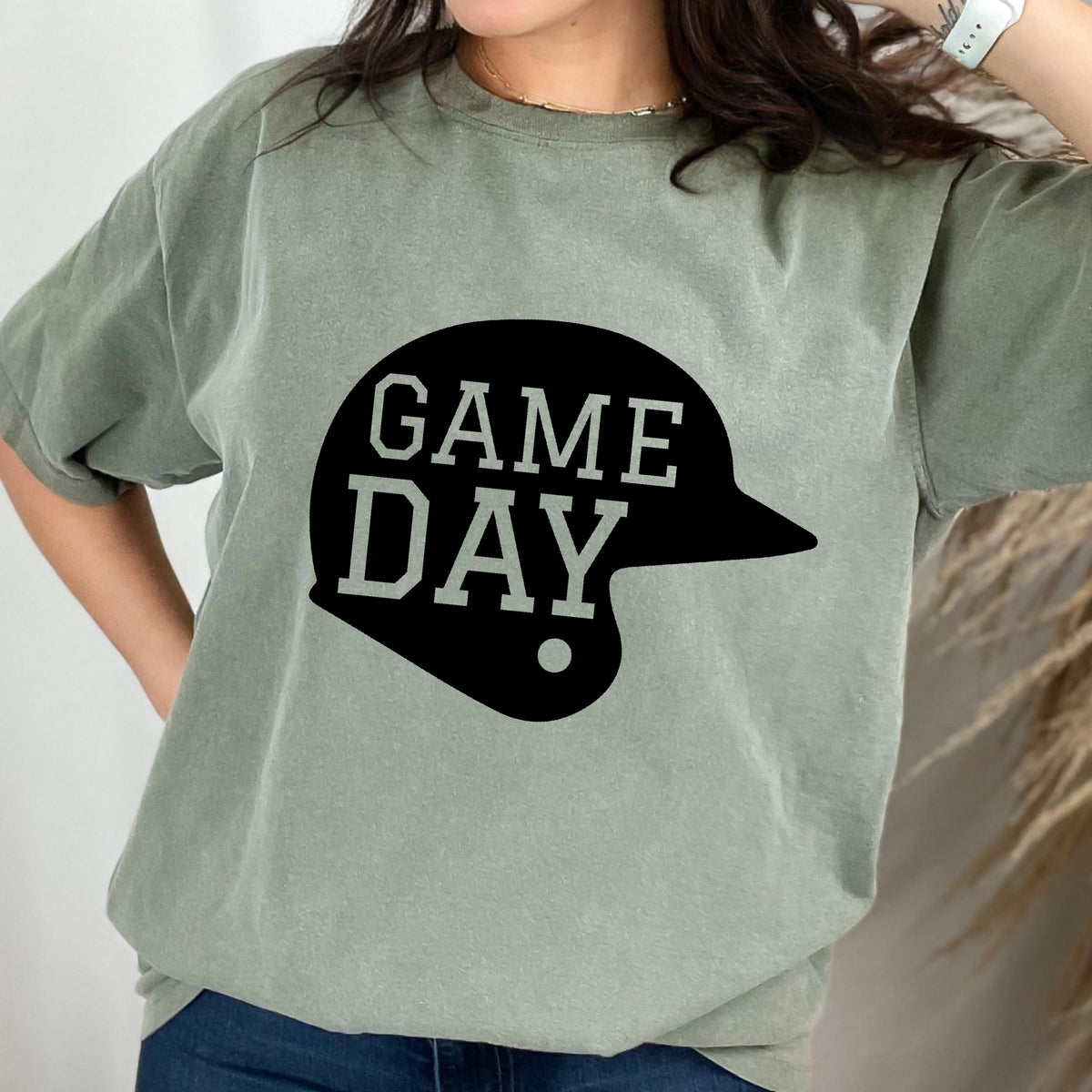 May include: A sage green short-sleeve t-shirt featuring a black graphic of a baseball helmet silhouette with the text GAME DAY printed in a bold, block-style font across the center of the helmet design.