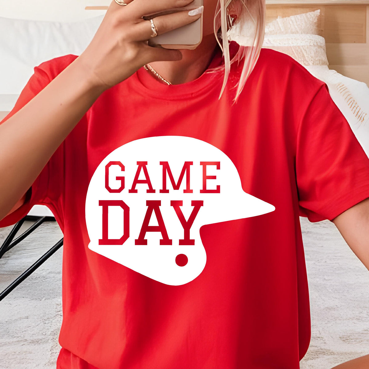 May include: A bright red short-sleeved t-shirt featuring a large white graphic of a baseball helmet silhouette. Inside the helmet shape, the text "GAME DAY" is printed in bold red capital letters. The shirt has a classic crew neckline.