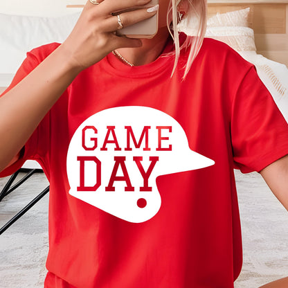 May include: A bright red short-sleeved t-shirt featuring a large white graphic of a baseball helmet silhouette. Inside the helmet shape, the text "GAME DAY" is printed in bold red capital letters. The shirt has a classic crew neckline.