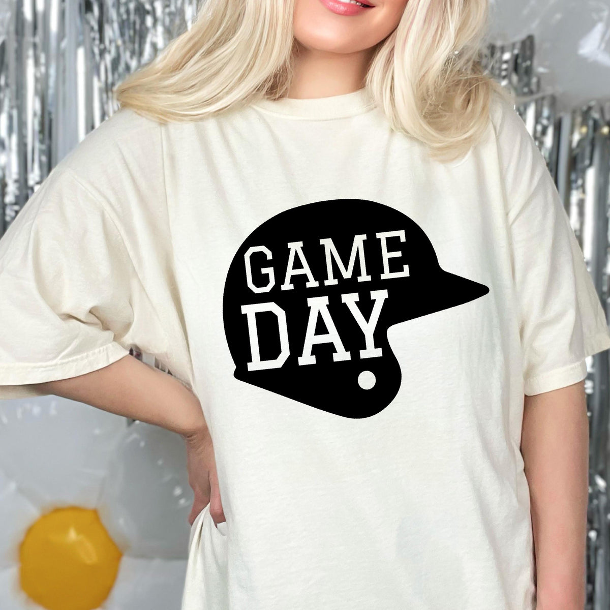 May include: A cream-colored short-sleeve t-shirt featuring a large black graphic of a baseball helmet silhouette. The text GAME DAY is printed in white, bold, block-style lettering across the center of the black helmet graphic.