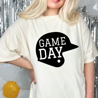 May include: A cream-colored short-sleeve t-shirt featuring a large black graphic of a baseball helmet silhouette. The text GAME DAY is printed in white, bold, block-style lettering across the center of the black helmet graphic.
