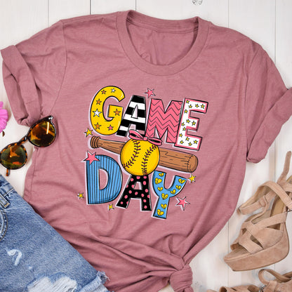 May include: A pink t-shirt with the words "GAME DAY" in colorful letters with a baseball and bat graphic. The letters are decorated with stars, hearts, and polka dots.