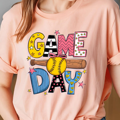 May include: A pink t-shirt with a colorful graphic design that says "GAME DAY" with a baseball and bat. The letters are decorated with stars, hearts, and polka dots.