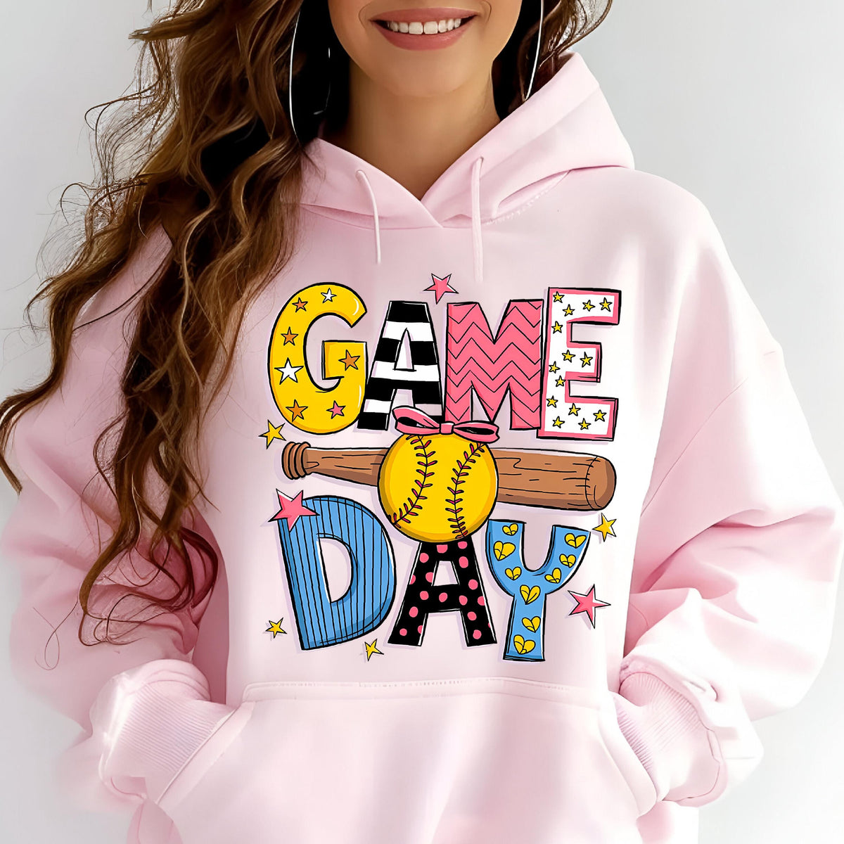 May include: A light pink hooded sweatshirt featuring a graphic print with the text GAME DAY in colorful, patterned letters. A brown baseball bat and a yellow softball with a pink bow are centered in the design, surrounded by small pink stars.