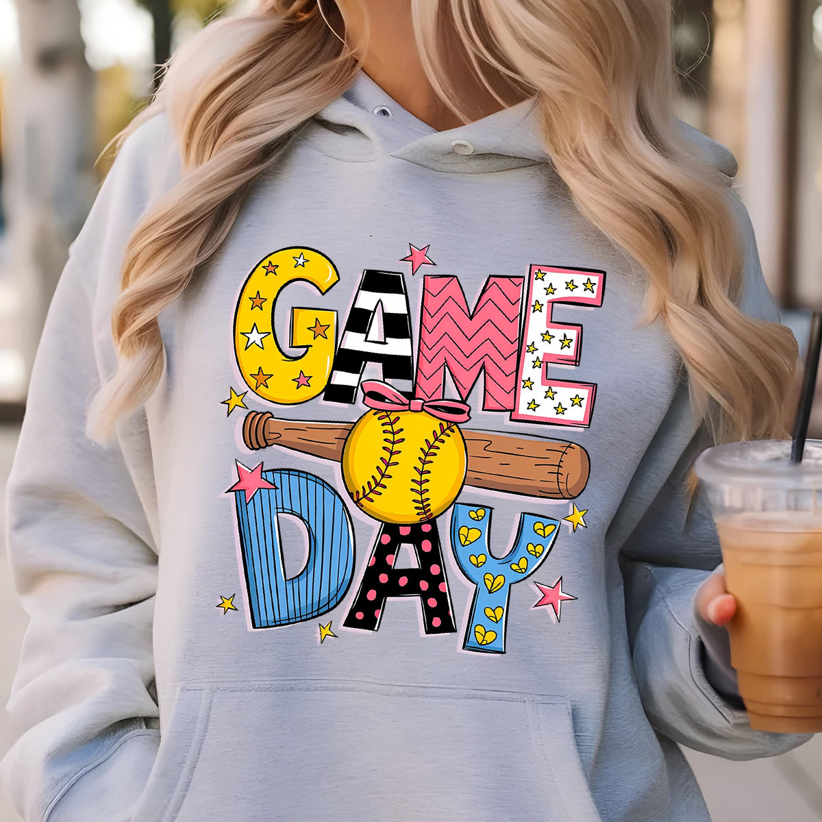 May include: A light gray hooded sweatshirt featuring a graphic print with the text GAME DAY. The design includes a yellow softball with a pink bow, a brown wooden baseball bat, and various colorful patterns including stars, stripes, and dots.