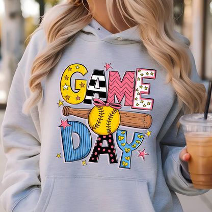 May include: A light gray hooded sweatshirt featuring a graphic print with the text GAME DAY. The design includes a yellow softball with a pink bow, a brown wooden baseball bat, and various colorful patterns including stars, stripes, and dots.