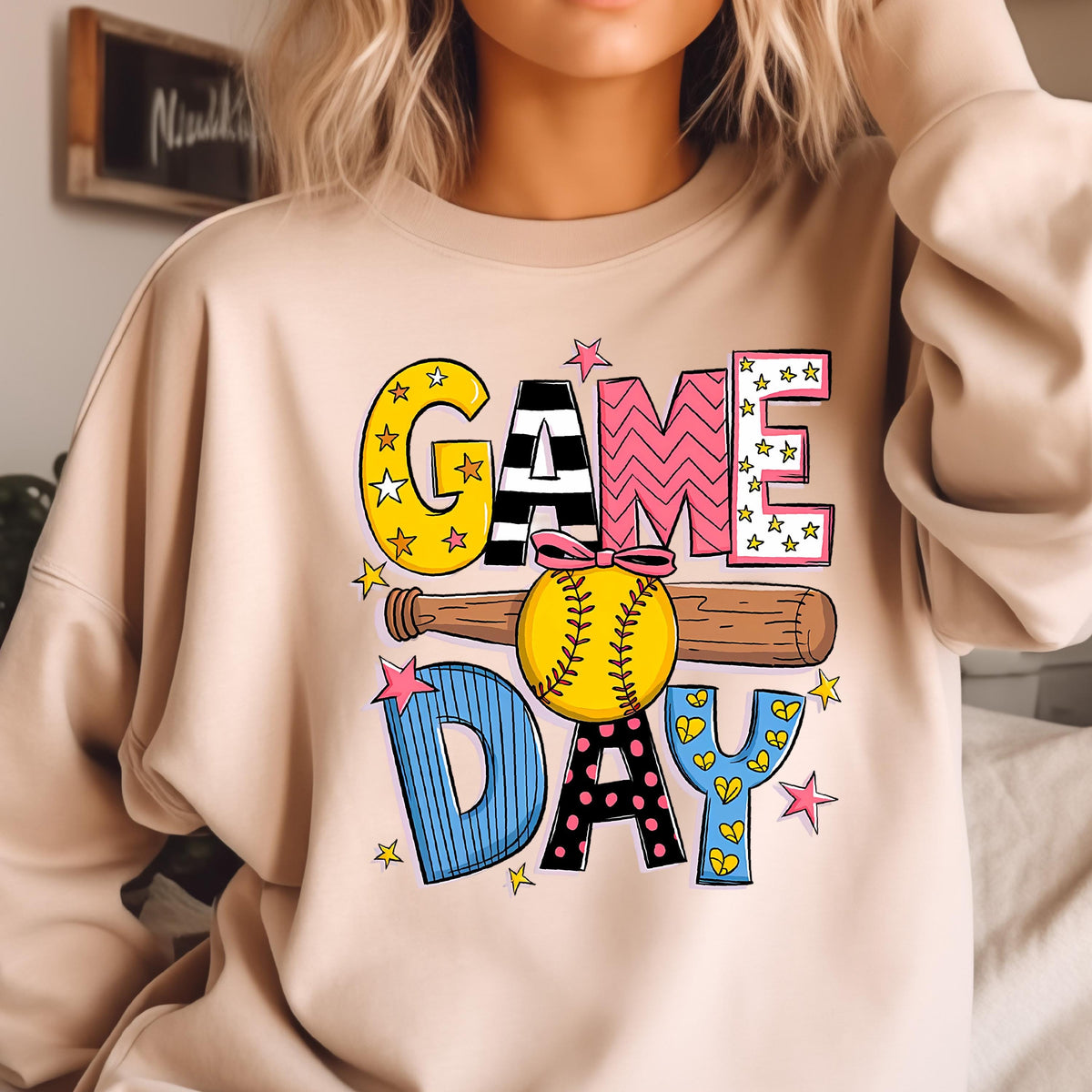 May include: A beige crewneck sweatshirt featuring a graphic print with the text "GAME DAY". The design includes a yellow softball with a pink bow, a brown baseball bat, and colorful letters decorated with stars, stripes, and polka dots.