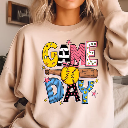 May include: A beige crewneck sweatshirt featuring a graphic print with the text "GAME DAY". The design includes a yellow softball with a pink bow, a brown baseball bat, and colorful letters decorated with stars, stripes, and polka dots.