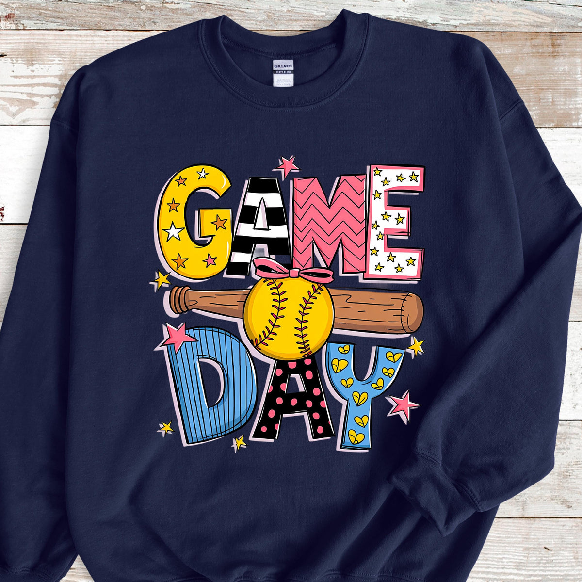 May include: A navy blue crewneck sweatshirt featuring a graphic print with the text GAME DAY. The letters are decorated with yellow stars, black and white stripes, pink chevrons, blue pinstripes, and polka dots, centered with a softball and bat.
