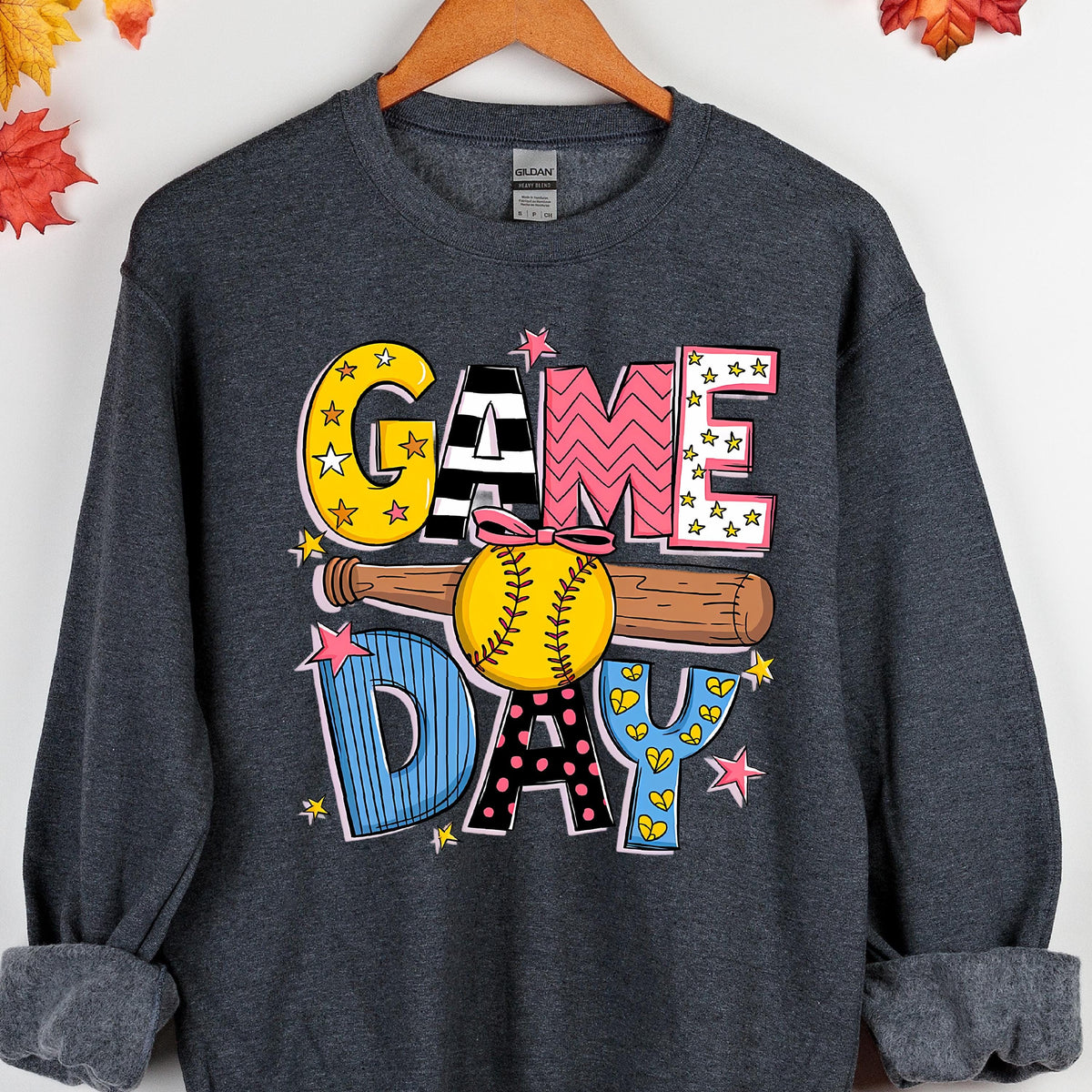 May include: A dark heather gray long-sleeve sweatshirt featuring a graphic print with the text GAME DAY. The design includes a brown baseball bat and a yellow softball with a pink bow, surrounded by colorful stars and patterned letters.