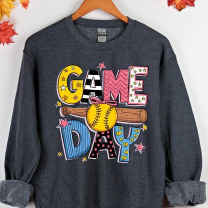 May include: A dark heather gray long-sleeve sweatshirt featuring a graphic print with the text GAME DAY. The design includes a brown baseball bat and a yellow softball with a pink bow, surrounded by colorful stars and patterned letters.