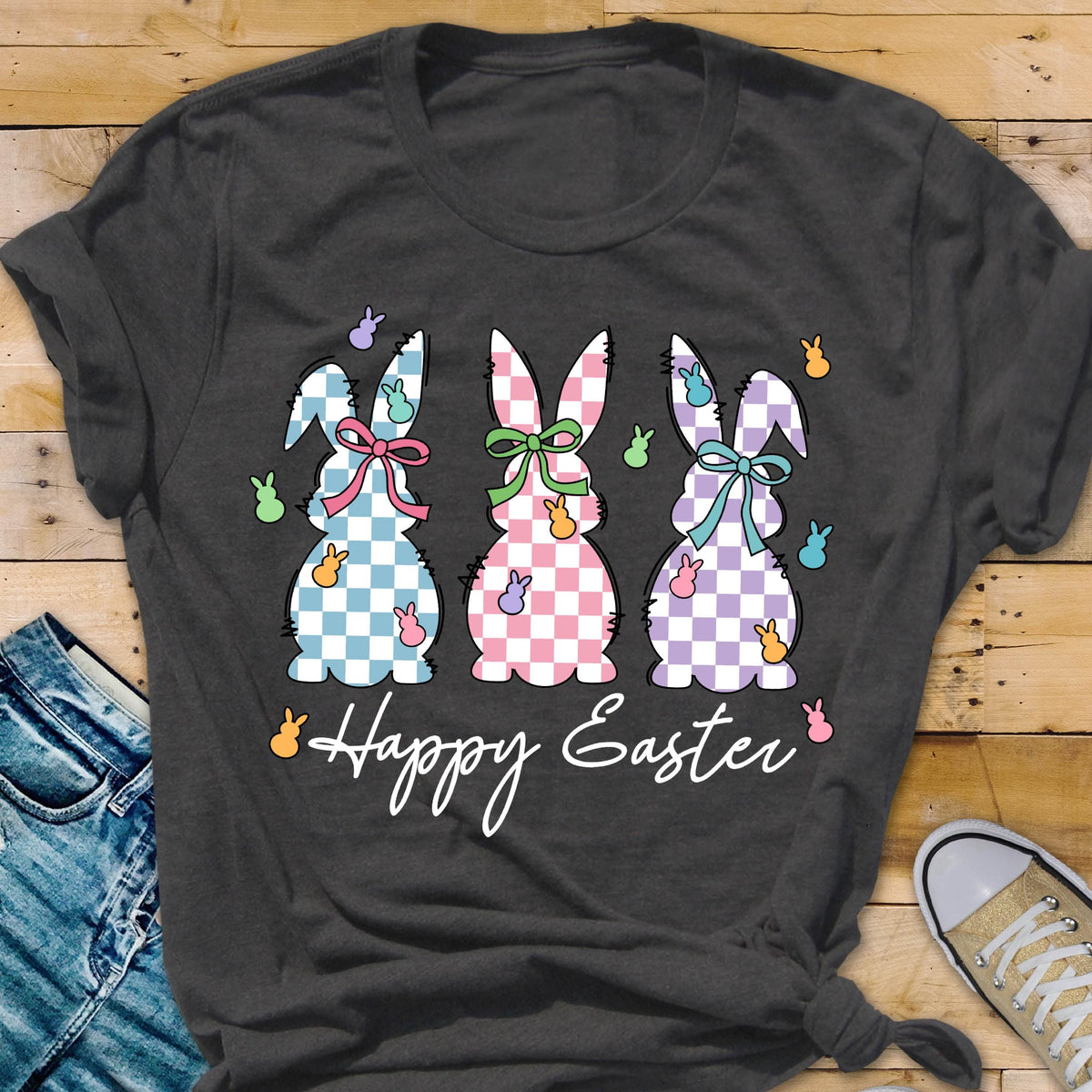 May include: A dark gray t-shirt with a graphic of three checkered Easter bunnies in blue, pink, and purple. The bunnies have bows and are surrounded by small, colorful Easter eggs. The text "Happy Easter" is printed below the bunnies.