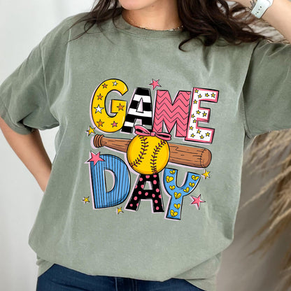 May include: A green t-shirt with a colorful graphic design that says "GAME DAY" with a baseball and bat. The graphic is in a cartoon style with stars, hearts, and polka dots.