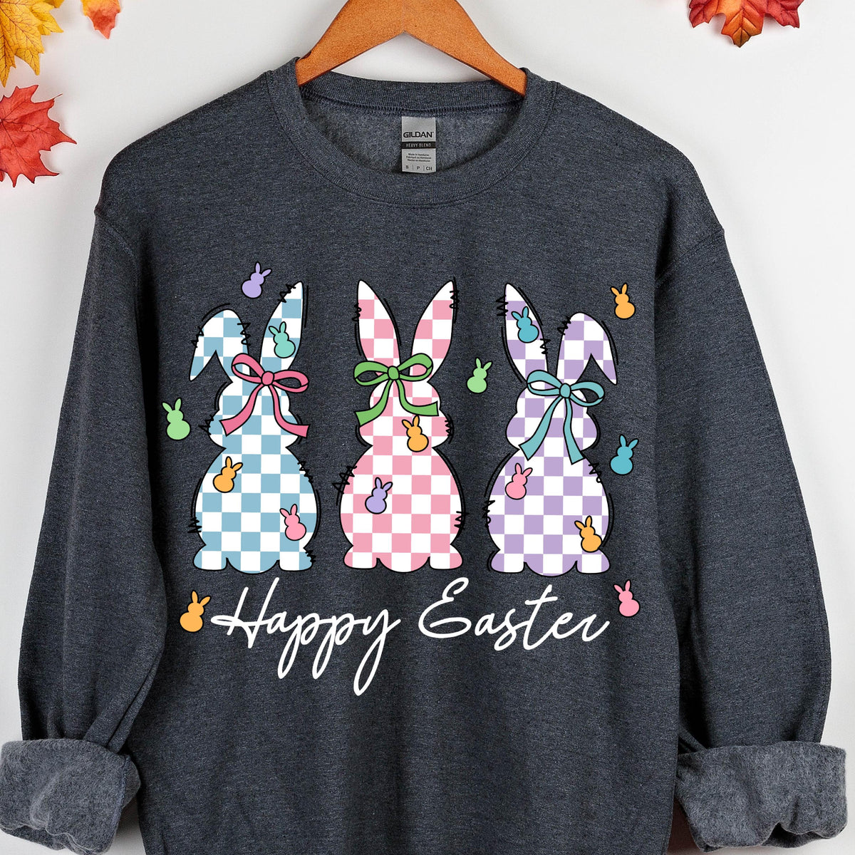 May include: A dark gray sweatshirt with a graphic of three Easter bunnies in a row. The bunnies are wearing checkered patterns in blue, pink, and purple. The text "Happy Easter" is printed below the bunnies.