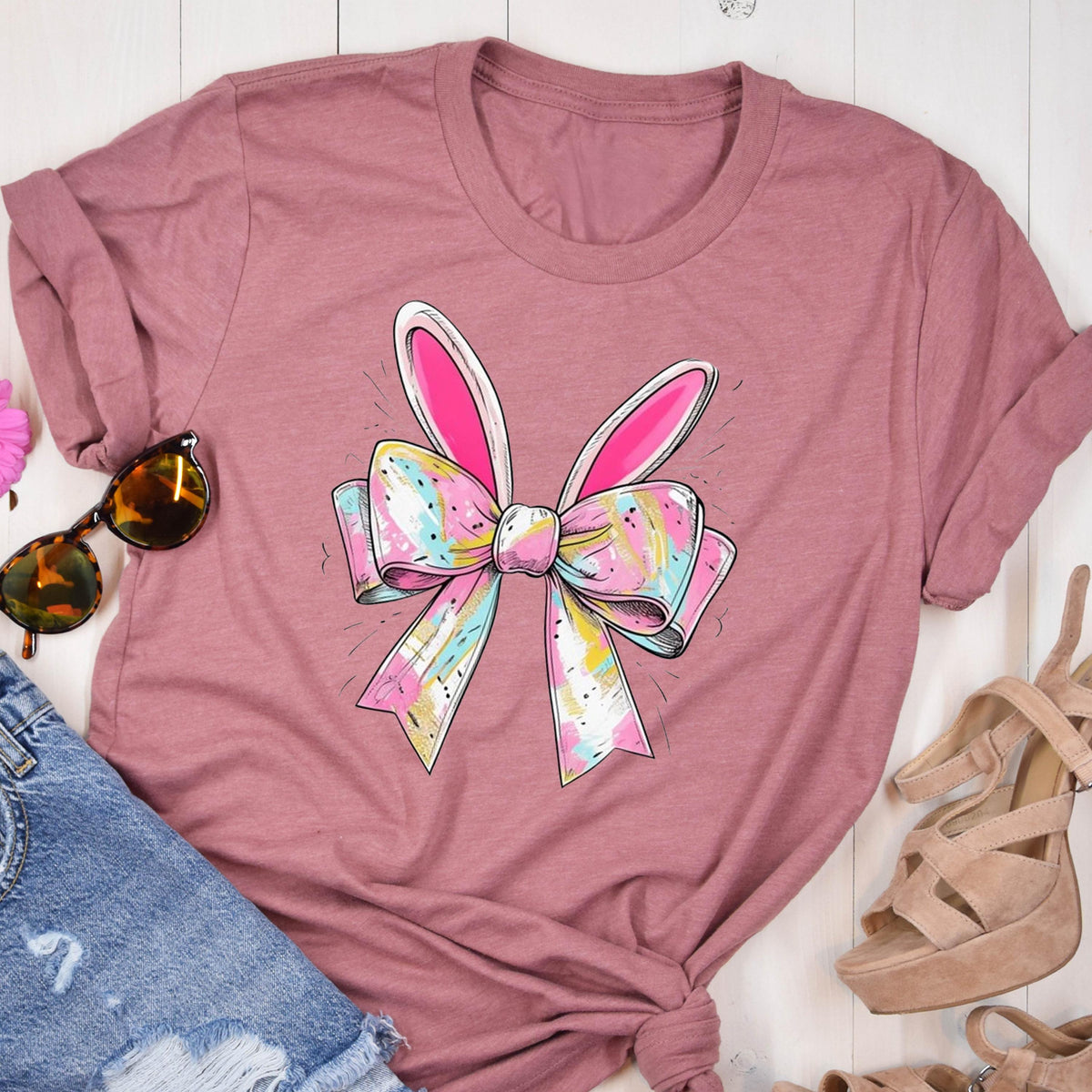 May include: A pink t-shirt with a colorful bow and bunny ears design. The bow is made of pastel colors and has a white outline. The bunny ears are pink and white.