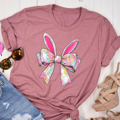May include: A pink t-shirt with a colorful bow and bunny ears design. The bow is made of pastel colors and has a white outline. The bunny ears are pink and white.