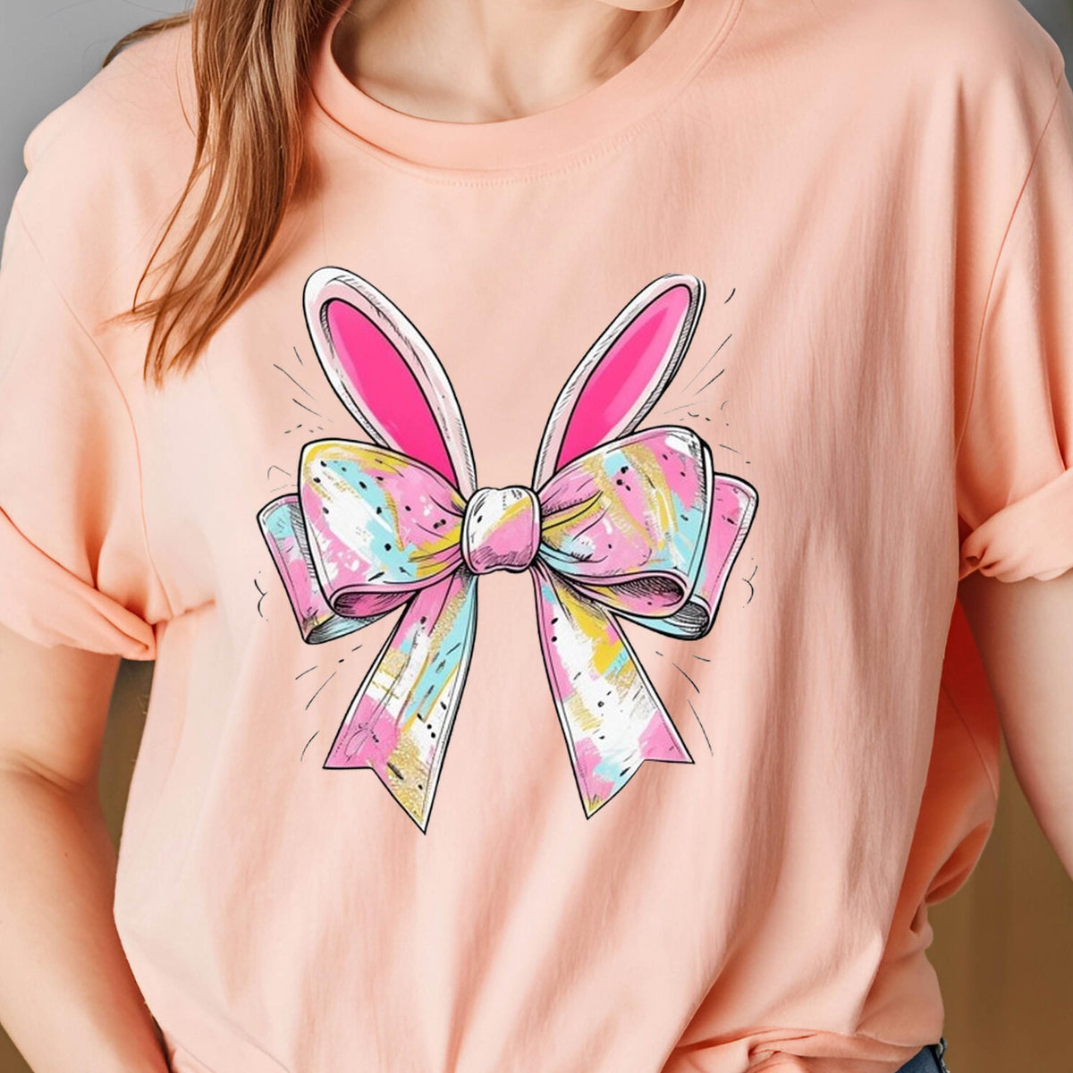May include: A pink t-shirt with a colorful bow and bunny ears design. The bow is made of pink, blue, white, and gold fabric.