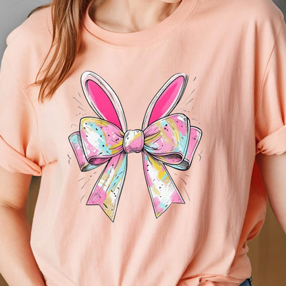 May include: A pink t-shirt with a colorful bow and bunny ears design. The bow is made of pink, blue, white, and gold fabric.