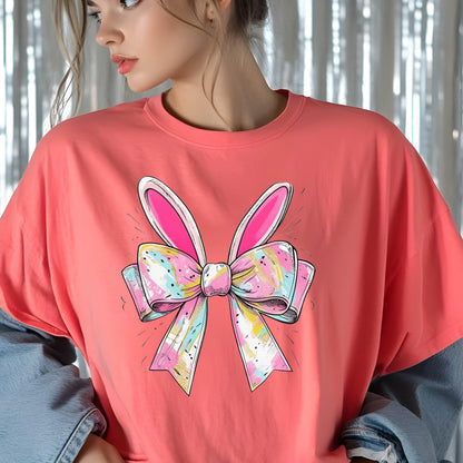 May include: A coral pink t-shirt with a graphic of a pink bow with bunny ears. The bow is decorated with colorful swirls and dots.