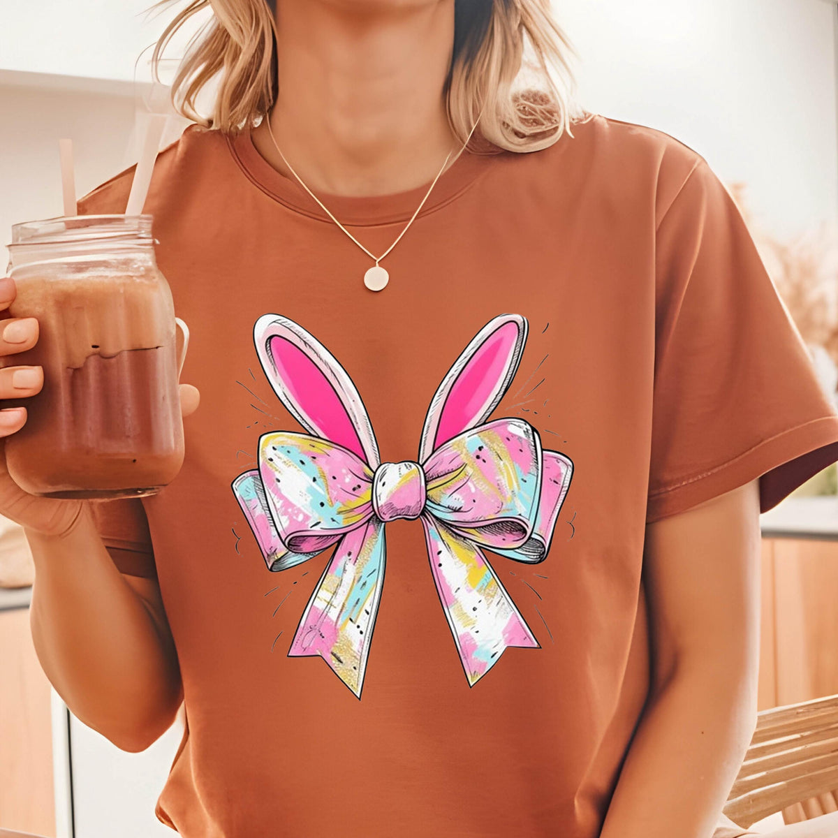 May include: A brown t-shirt with a colorful bow and bunny ears graphic. The bow is pink, yellow, blue, and white.
