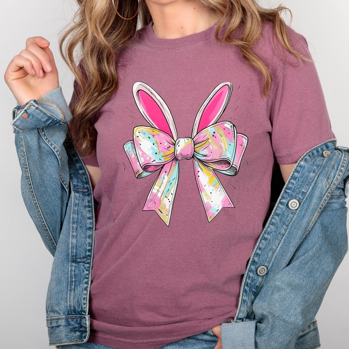 May include: A pink t-shirt with a colorful bow and bunny ears. The bow is made of pink, blue, and yellow ribbons with white accents. The bunny ears are white with pink insides.