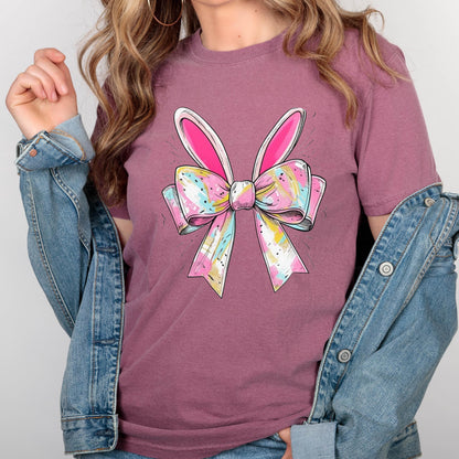 May include: A pink t-shirt with a colorful bow and bunny ears. The bow is made of pink, blue, and yellow ribbons with white accents. The bunny ears are white with pink insides.