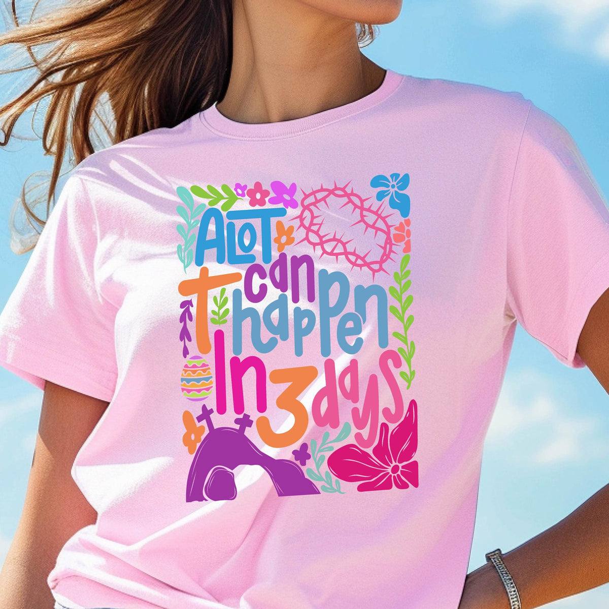 May include: Pink t-shirt with a colorful graphic design that says "A lot can happen in 3 days". The design includes flowers, a crown of thorns, and a tomb.