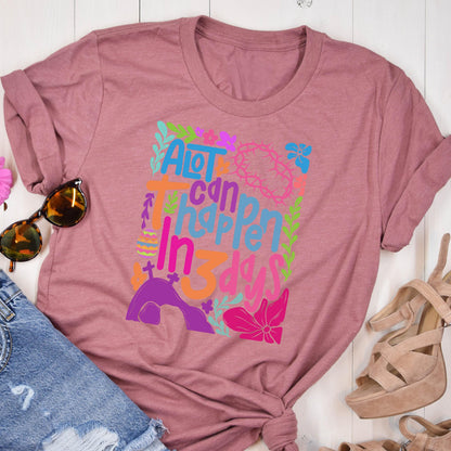 May include: A pink t-shirt with a colorful graphic design that says "A lot can happen in 3 days". The graphic includes flowers, leaves, and a cross.