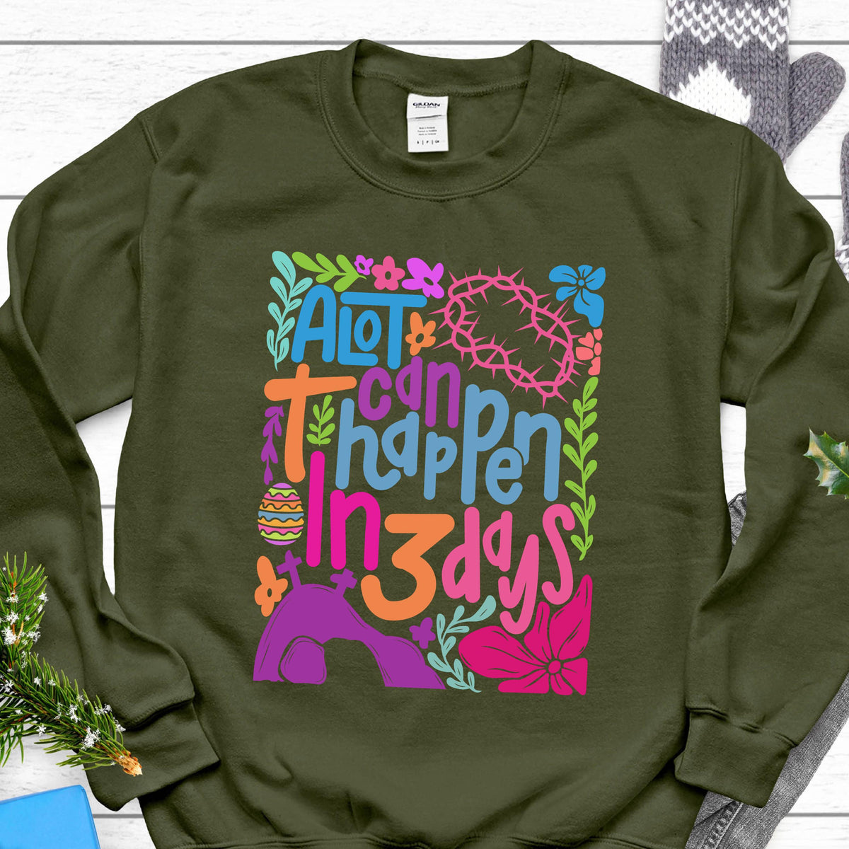 May include: A green sweatshirt with a colorful graphic design that says "A lot can happen in 3 days". The graphic includes flowers, a crown of thorns, and an Easter egg.