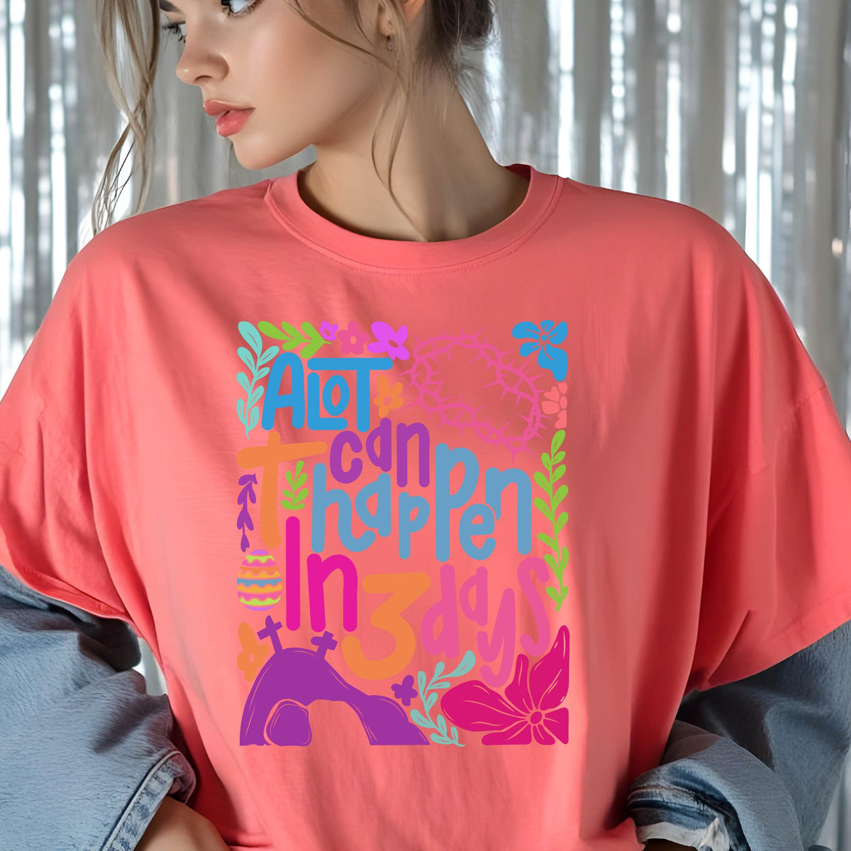 May include: A pink t-shirt with a colorful graphic design that says "A lot can happen in 3 days". The graphic includes flowers, a cross, a crown of thorns, and an Easter egg.