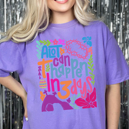 May include: Purple t-shirt with a colorful graphic design that says "A lot can happen in 3 days". The graphic includes a cross, a crown of thorns, an Easter egg, and a tomb.