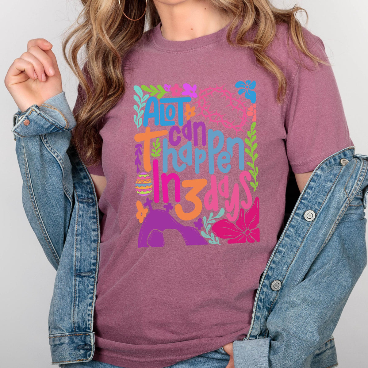 May include: A purple t-shirt with a colorful graphic design that says "A lot can happen in 3 days". The graphic includes flowers, leaves, and an Easter egg.