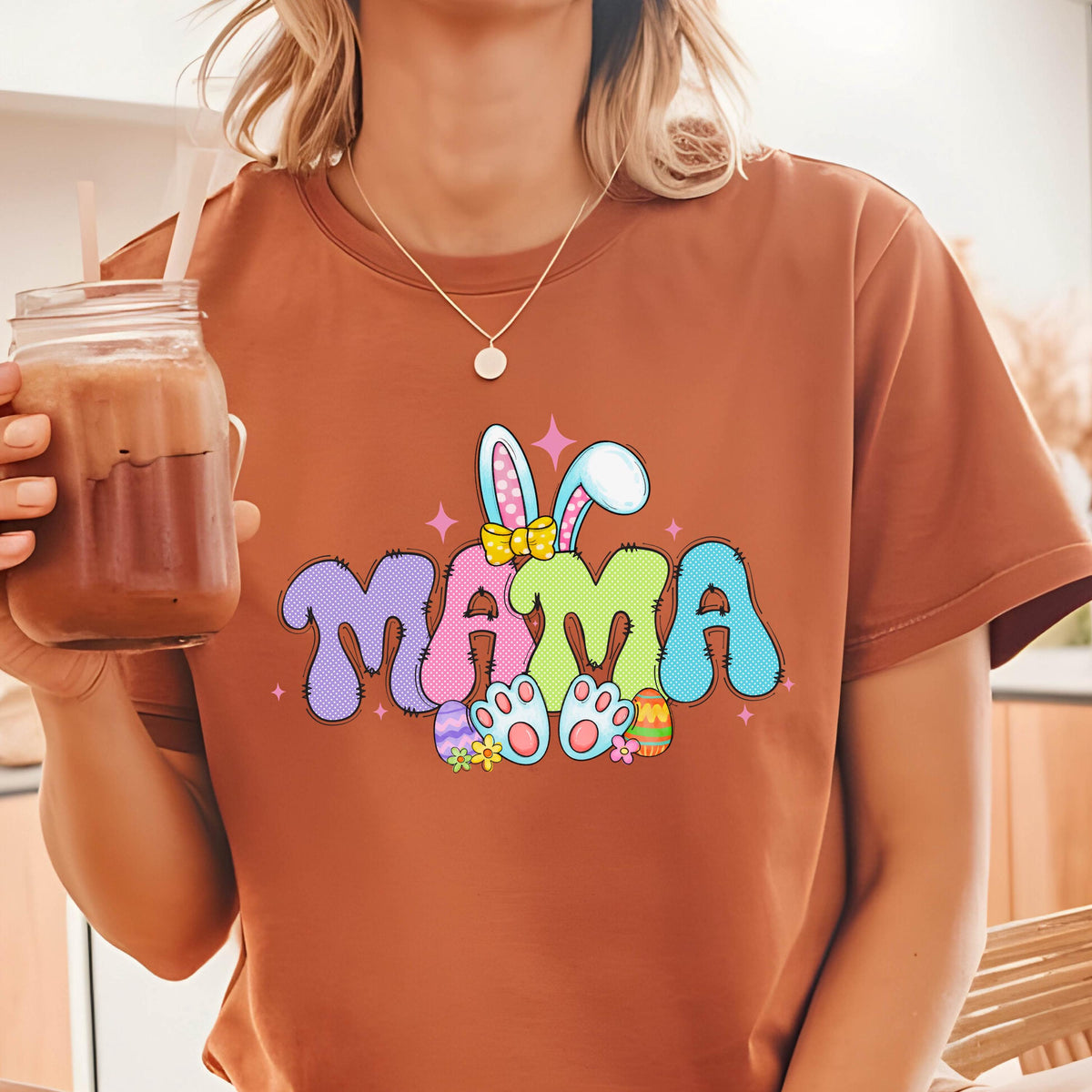 May include: A rust-colored t-shirt featuring the word "MAMA" in a colorful, whimsical design. The design includes bunny ears, Easter eggs, and flower accents, perfect for Easter or Mother's Day.