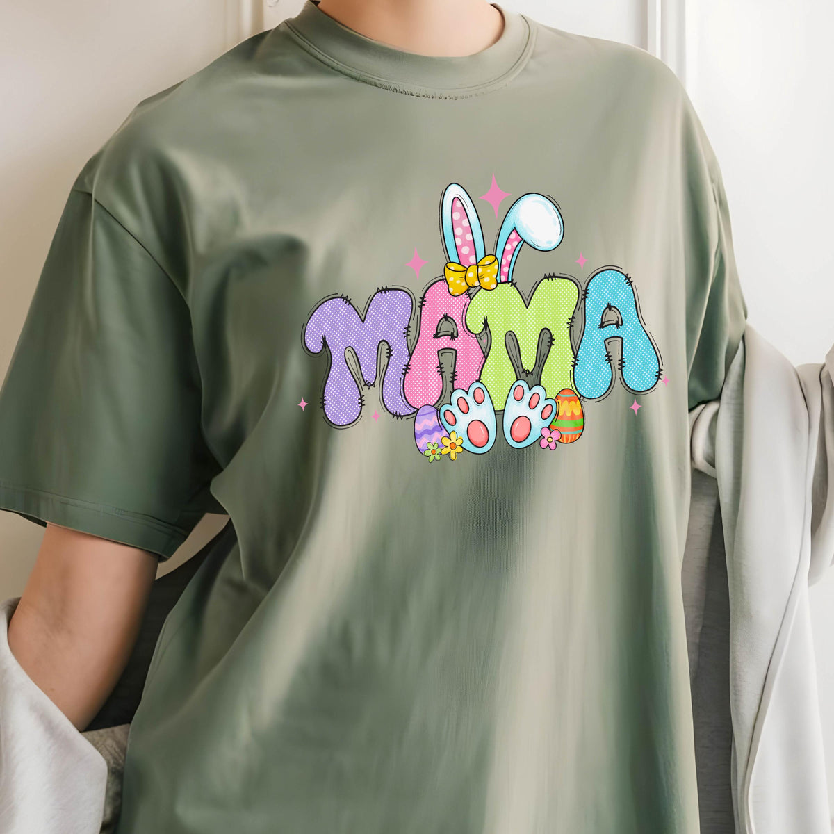 May include: Olive green t-shirt featuring the word "MAMA" in a colorful, bubble letter design with bunny ears and Easter eggs. The graphic includes pink, purple, blue, yellow, and green colors. A casual, short-sleeved tee.