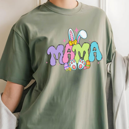 May include: Olive green t-shirt featuring the word "MAMA" in a colorful, bubble letter design with bunny ears and Easter eggs. The graphic includes pink, purple, blue, yellow, and green colors. A casual, short-sleeved tee.