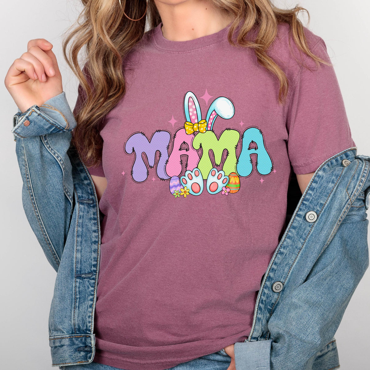 May include: A mauve-colored t-shirt with the word "MAMA" in colorful, bubble letters. The design includes bunny ears, Easter eggs, and bunny feet, creating a festive Easter theme. The shirt is a short-sleeved crew neck.